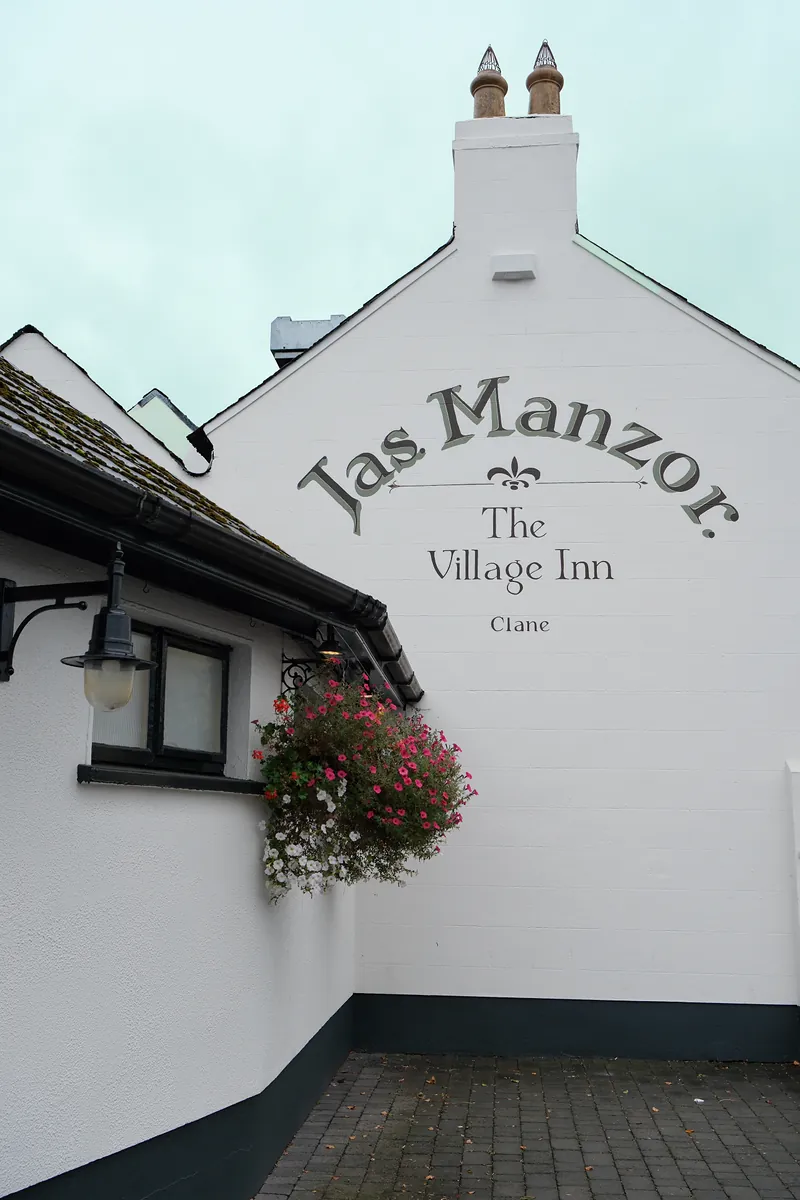 The painted Jas. Manzor. The Village Inn, Clane sign on the white gable wall, with a hanging basket of pink flowers