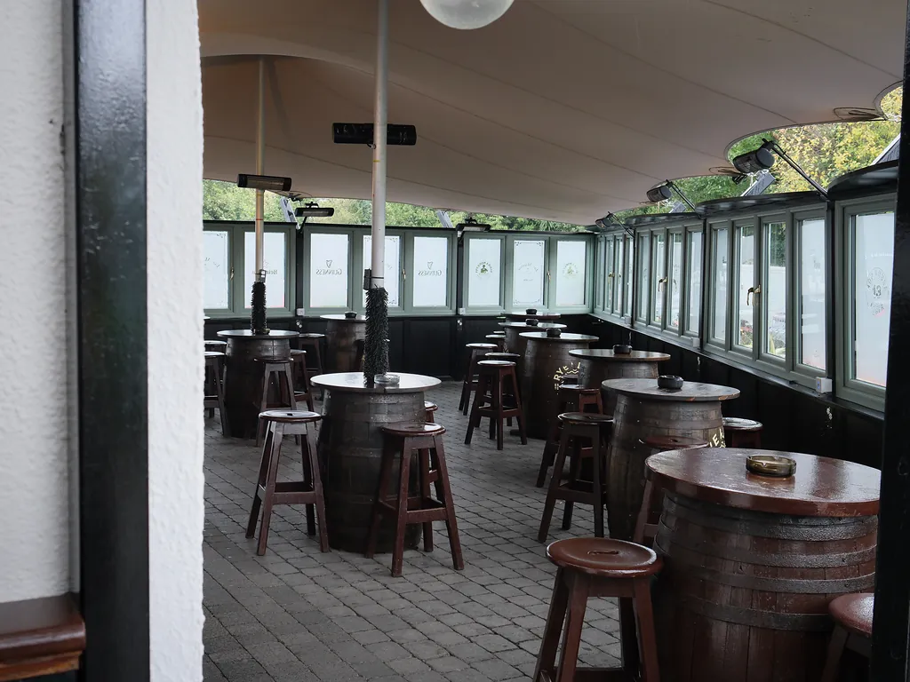 The covered beer garden: barrel tables and wooden stools beneath a white canopy, with heaters overhead
