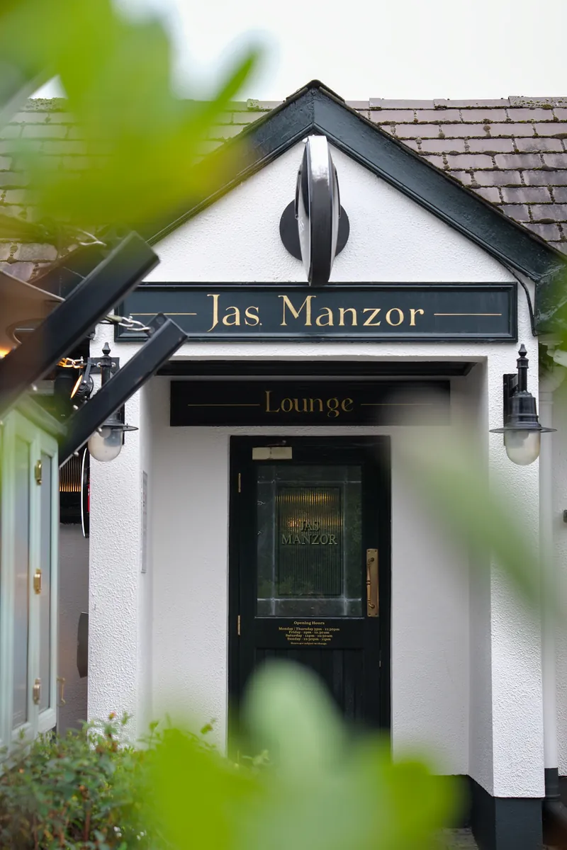 The Jas Manzor Lounge entrance seen through green leaves, a white porch with a dark green door