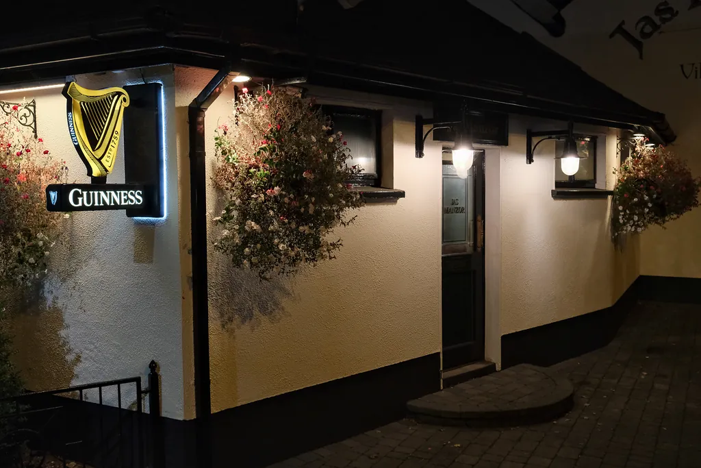 A Jas Manzor doorway at night, a glowing Guinness harp sign and hanging baskets lit by lanterns